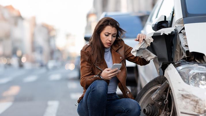 Mujer joven contactando con su seguro por el móvil tras sufrir un accidente de coche en carretera, sentada en el arcén junto a su vehículo con daños visibles